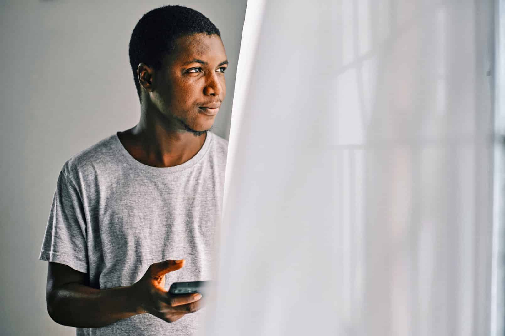 Man with cell phone in hand looks out of window, blowing white curtain in foreground